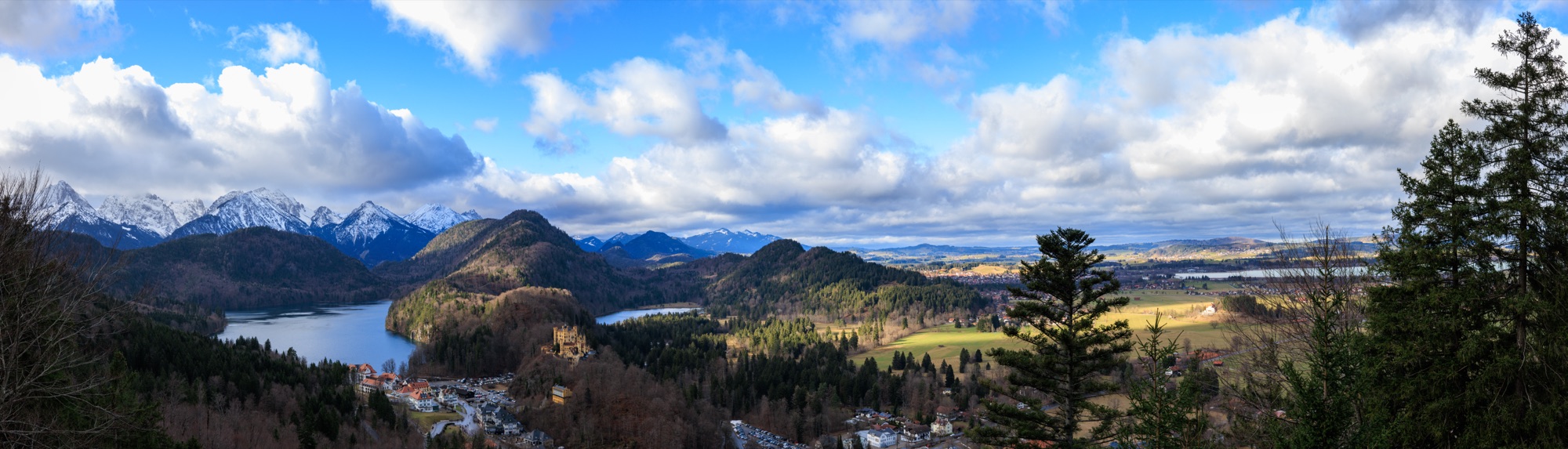 Alpenpanorama mit See, bewaldeten Hügeln und schneebedeckten Gipfeln unter Wolkenhimmel