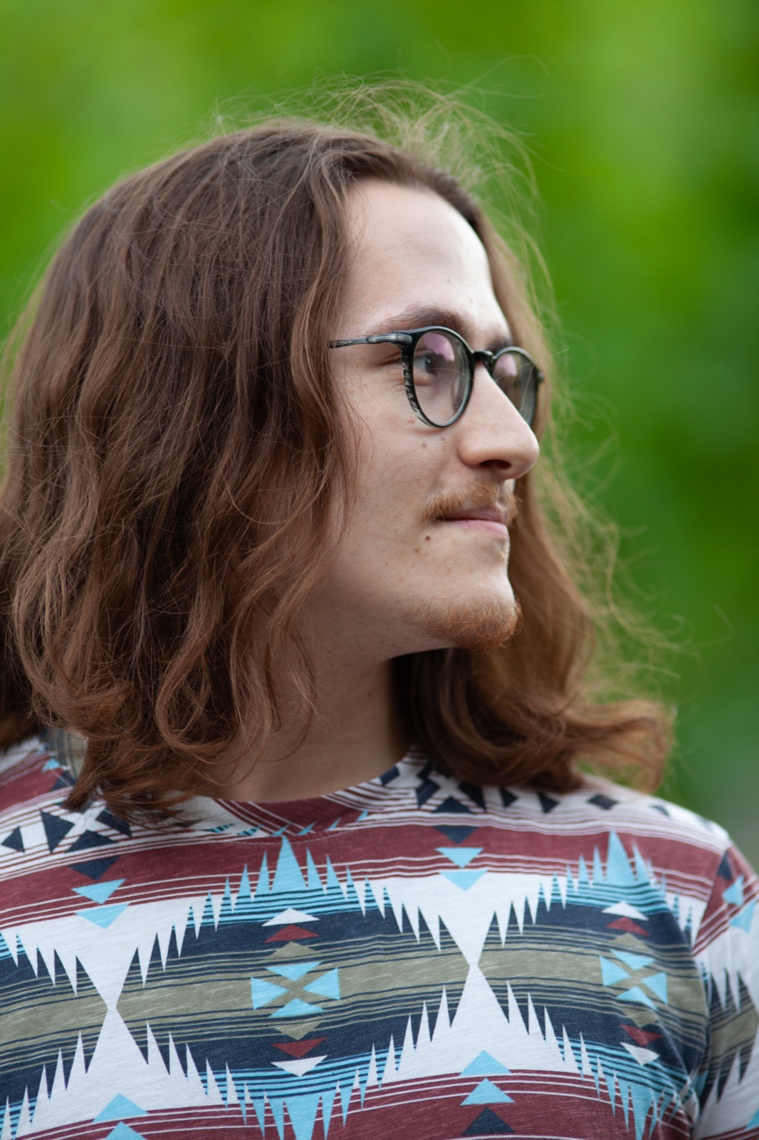 Porträt eines jungen Mannes mit langen Haaren und runder Brille im Profil vor grünem Hintergrund